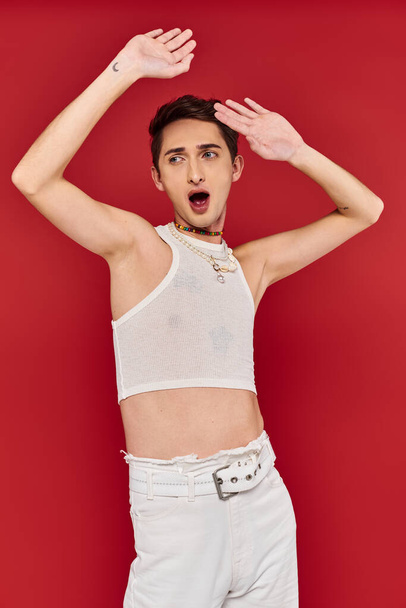 шокированный модный гей в повседневной одежде с аксессуарами на красном фоне и глядя в сторону - Фото, изображение