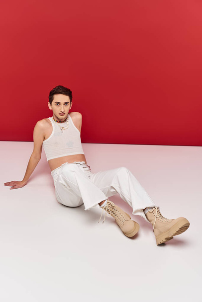 gut aussehender androgyner Mann in lässiger Kleidung, der auf dem Boden sitzt und vor rotem Hintergrund in die Kamera blickt - Foto, Bild