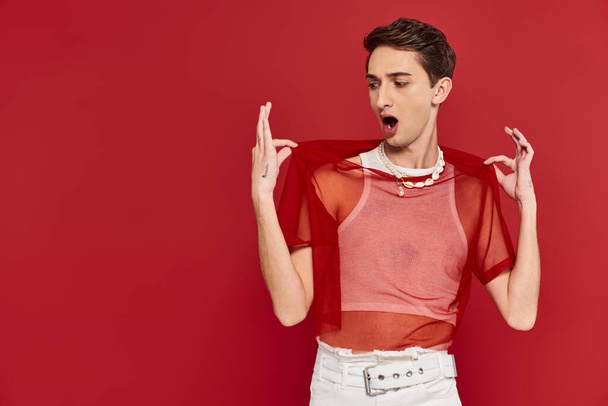 handsome shocked androgynous man with red stylish fishnet posing emotionally on red backdrop - Photo, Image