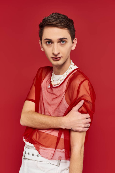 attractive voguish androgynous man with stylish red fishnet looking at camera on red backdrop - Foto, afbeelding