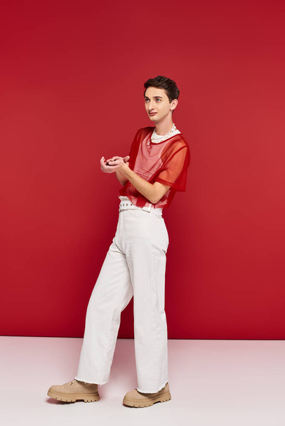 pohledný androgynní muž v bílém oblečení s červenou síťkou pózující na červeném pozadí a odvracející pohled - Fotografie, Obrázek
