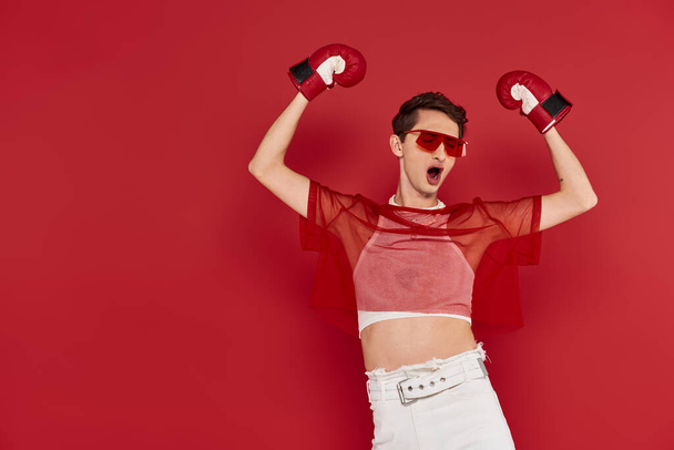 alegre hombre andrógino de moda con red roja posando con guantes de boxeo con los ojos cerrados - Foto, imagen