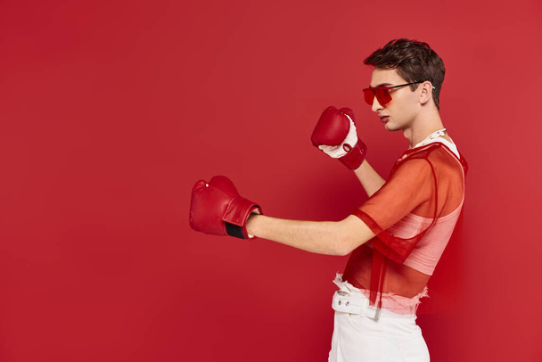 fashionable androgynous man with red fishnet posing actively with boxing gloves and looking away - Photo, Image