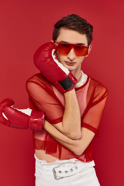 appealing gay man with sunglasses and red fishnet posing with boxing gloves and looking at camera - Fotó, kép