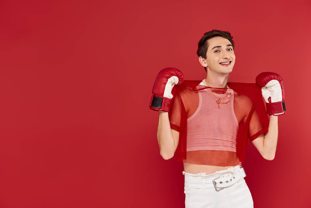 fröhlicher gut gekleideter schwuler Mann in lässiger Kleidung mit Boxhandschuhen vor rotem Hintergrund, der in die Kamera blickt - Foto, Bild