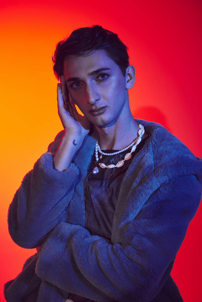 elegant androgynous man in purple faux fur posing on vibrant backdrop and looking at camera - Фото, изображение