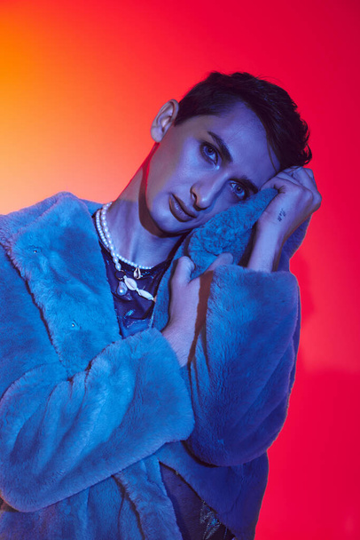 sophisticated androgynous man in purple faux fur posing on vibrant backdrop and looking at camera - Fotó, kép