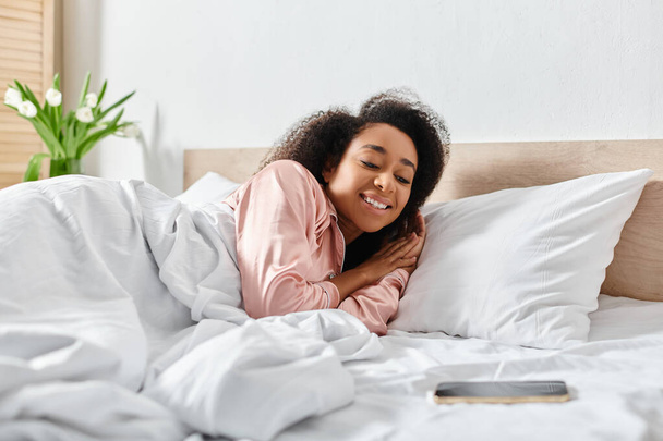 Lockige Afroamerikanerin im Schlafanzug lehnt sich friedlich auf einem Bett mit weißen Laken in einem gemütlichen Schlafzimmer. - Foto, Bild