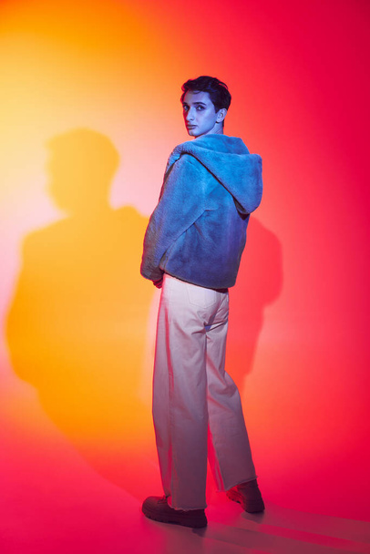 well-dressed androgynous man in purple faux fur posing on vibrant backdrop and looking at camera - Photo, Image
