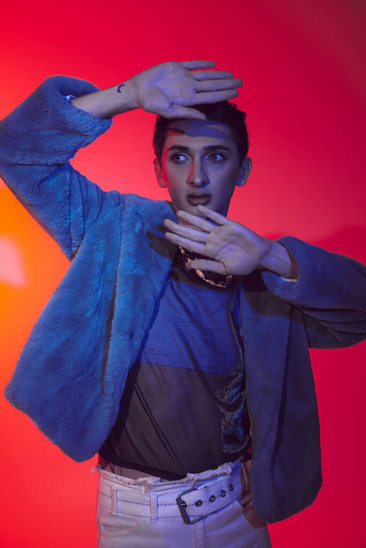 attractive modish androgynous man in purple faux fur posing on vibrant backdrop and looking away - Foto, Imagem