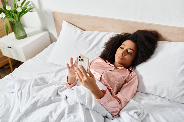 Σγουρή Αφρο-Αμερικανίδα γυναίκα με πιτζάμες χαλαρώνει στο κρεβάτι, κρατώντας το κινητό στο φως του πρωινού. - Φωτογραφία, εικόνα