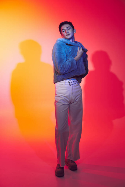fancy verleidelijke androgyne man in paars nepbont poseren met gesloten ogen op levendige achtergrond - Foto, afbeelding