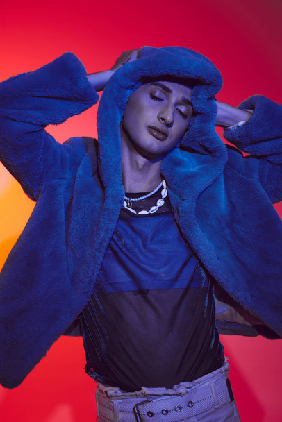 stylish alluring androgynous man in purple faux fur posing with closed eyes on vibrant backdrop - Photo, Image