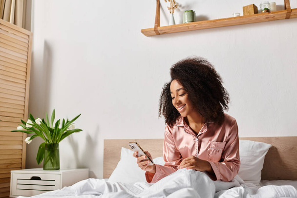 Riccio donna afroamericana in pigiama si siede sul letto, nel profondo del pensiero, concentrandosi sul suo schermo del telefono cellulare al mattino. - Foto, immagini