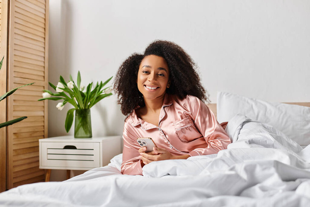 Lockige Afroamerikanerin im Schlafanzug sitzt auf dem Bett und hält morgens ihr Handy in der Hand. - Foto, Bild