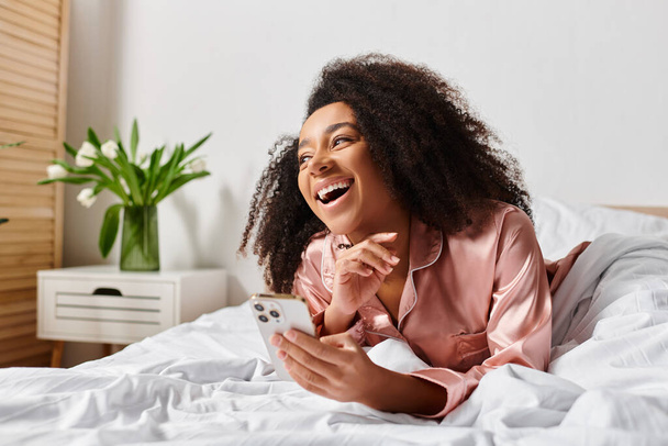 パジャマを着たカーリーなアフリカ系アメリカ人女性は,携帯電話を持っている間,平和的にベッドに横たわります.. - 写真・画像