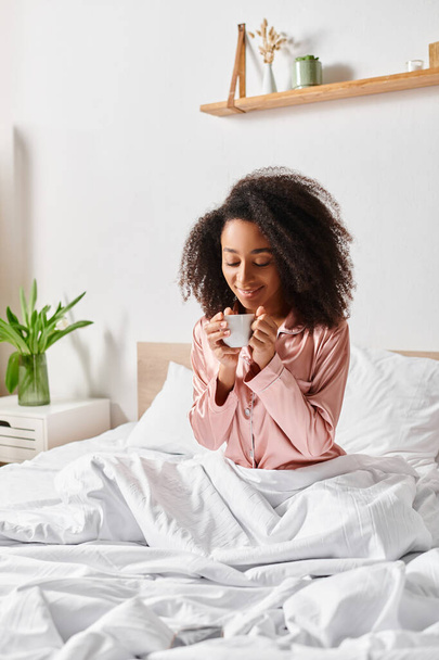 Egy göndör afro-amerikai nő pizsamában ül az ágyon, és reggelente nyugodtan kortyolgat egy csésze kávét.. - Fotó, kép
