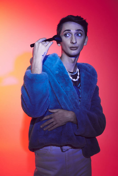 sophisticated androgynous man in stylish attire using makeup brush and posing on vibrant backdrop - Fotó, kép