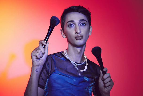elegant young androgynous man in casual attire holding two makeup brushes and looking at camera - Fotoğraf, Görsel
