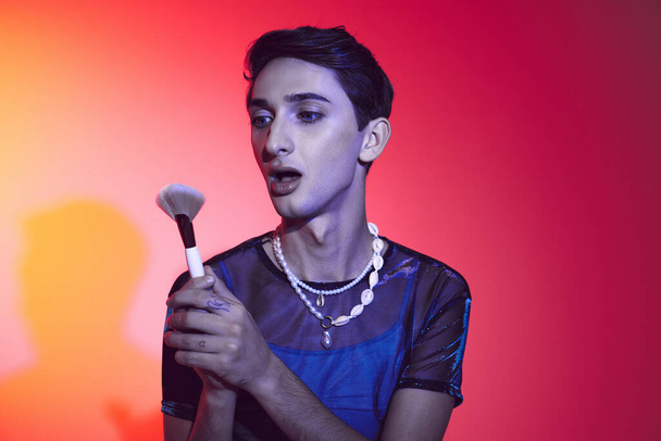 sophisticated androgynous man in stylish attire using makeup brush and posing on vibrant backdrop - Foto, Bild