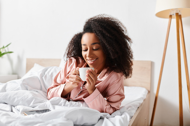 Kihara afroamerikkalainen nainen pyjamassa makaa sängyssä nauttien kupin kahvia aamuisin.. - Valokuva, kuva