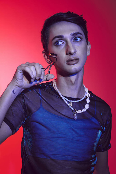 handsome fashionable androgynous man in casual attire holding eyelash curler on vibrant backdrop - Foto, Bild