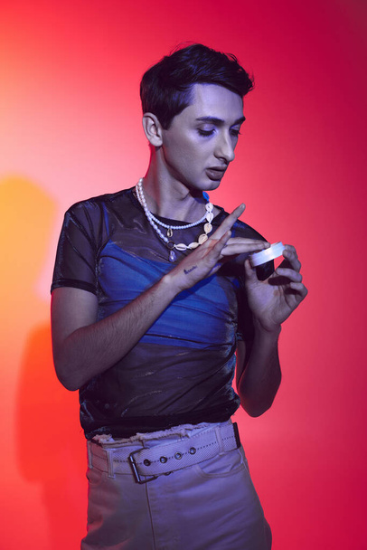κομψό όμορφος gay άντρας σε casual ζωντανή ενδυμασία κρατώντας κρέμα στα χέρια, ενώ σε πολύχρωμο φόντο - Φωτογραφία, εικόνα