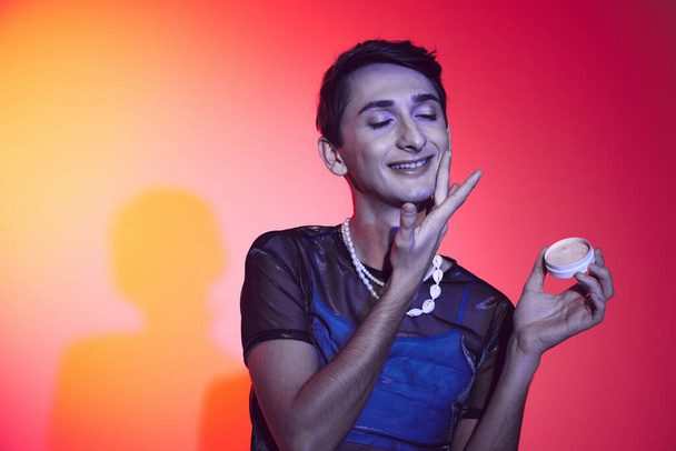 joyous handsome gay man in casual vibrant attire holding cream in hands while on colorful backdrop - Photo, Image
