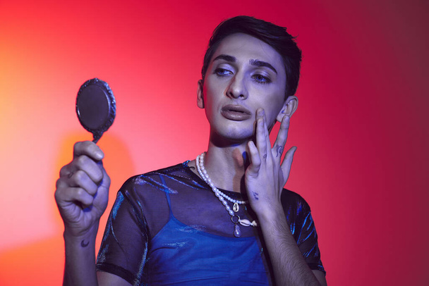 alluring androgynous man in vivid attire looking at small mirror while on red and orange backdrop - Foto, Bild