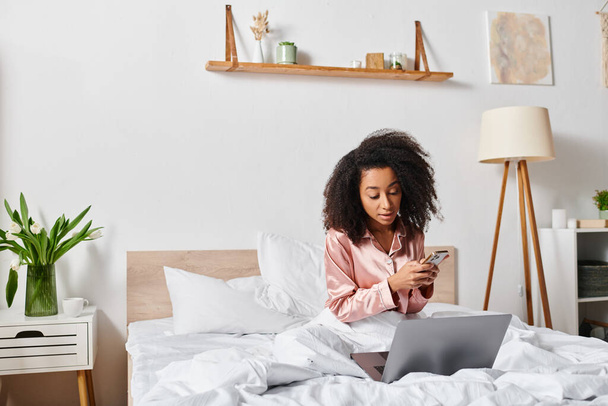Afrikkalainen amerikkalainen nainen pyjamassa istuu sängyllä, keskittyi kannettavan tietokoneen näytölle makuuhuoneessa aamulla. - Valokuva, kuva