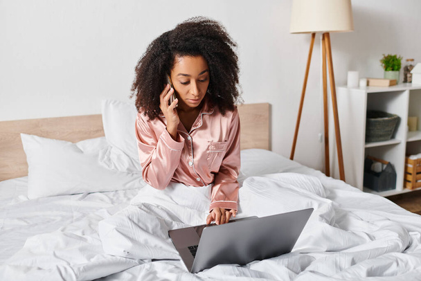 Pijamalı kıvırcık saçlı Afrikalı Amerikalı kadın yatakta oturur ve rahat yatak odasında dizüstü bilgisayar kullanmaya odaklanır.. - Fotoğraf, Görsel