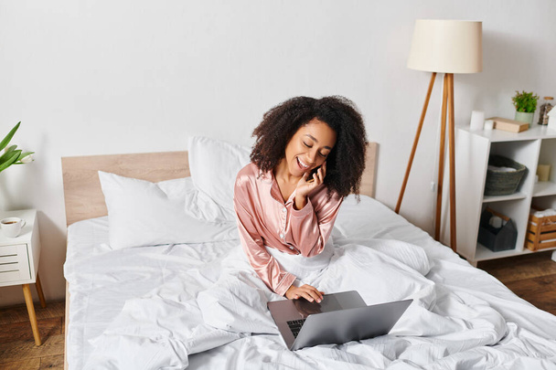 Göndör afro-amerikai nő pizsamában ül az ágyon, felszívódik a laptop képernyőjén, hangulatos hálószoba beállítás reggelente. - Fotó, kép