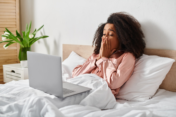 Een krullende Afro-Amerikaanse vrouw in pyjama ligt in bed met een laptop op haar schoot, bezig met technologie in een gezellige slaapkamer instelling. - Foto, afbeelding