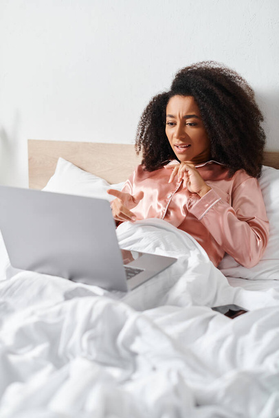 Yatakta pijamalı kıvırcık saçlı Amerikalı kadın sabahları yatak odasında dizüstü bilgisayar kullanmaya odaklanmış.. - Fotoğraf, Görsel