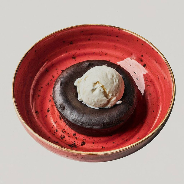 Een dessert, een chocoladetaart met een bolletje vanille-ijs, versierd met een muntblad, een eetbare bloem en een fysalis-vrucht, geserveerd op een rode keramische plaat.. - Foto, afbeelding