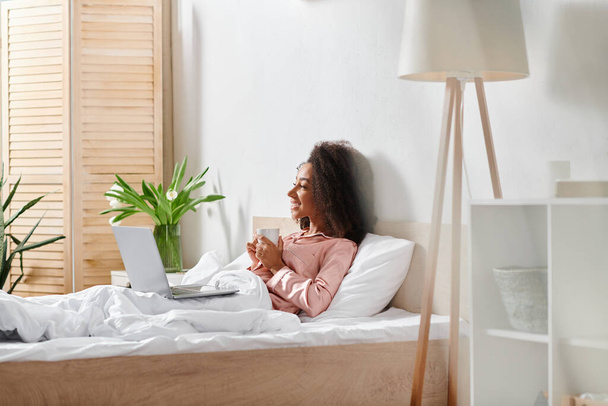 Kręcone Afroamerykanka kobieta w piżamie siedzi na łóżku, absorbowany w ekranie laptopa, rano światło filtrujące przez okno. - Zdjęcie, obraz