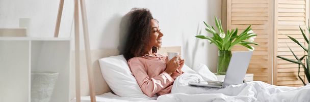 Σγουρή Αφροαμερικανή γυναίκα με πιτζάμες κάθεται στο κρεβάτι με ένα φορητό υπολογιστή. - Φωτογραφία, εικόνα
