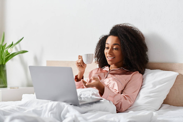 Mujer afroamericana rizada en pijama sentada en la cama, centrada en el uso de la computadora portátil en el acogedor dormitorio durante la mañana. - Foto, imagen