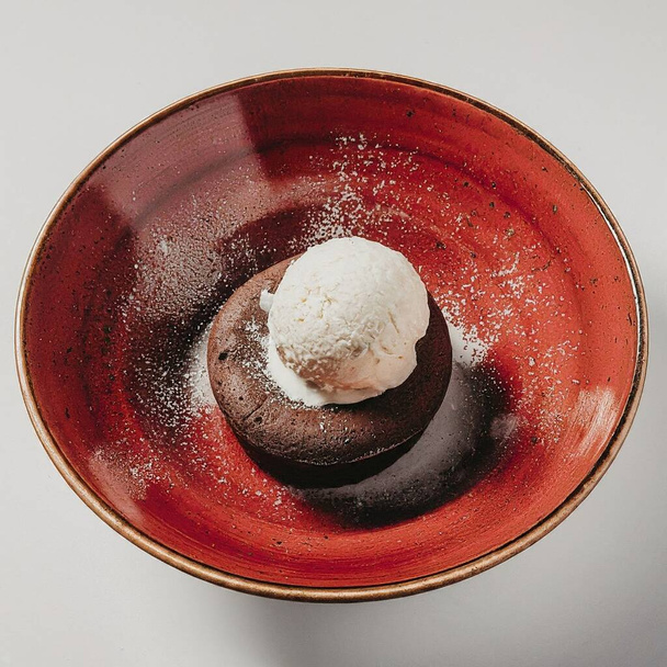 チョコレート溶岩ケーキはバニラアイスクリームのスクープでトッピングされ,ミントリーフ,食用花,そして物理的なフルーツで飾られ,すべてが光沢のある赤いセラミックプレートで提供されました. - 写真・画像