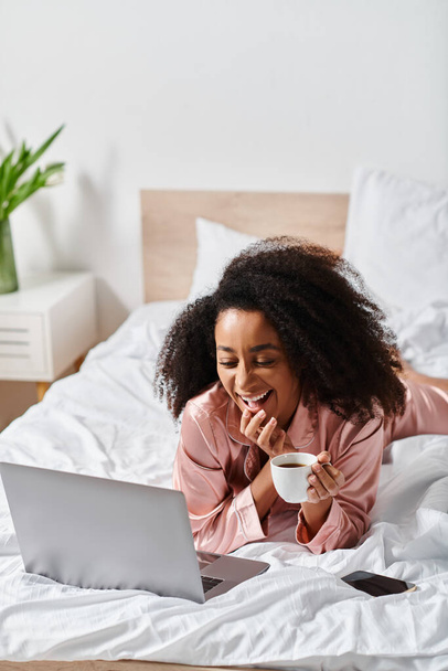 Kędzierzawa Afroamerykanka w piżamie spokojnie leży na łóżku, delektując się laptopem i filiżanką kawy w swojej sypialni rano. - Zdjęcie, obraz