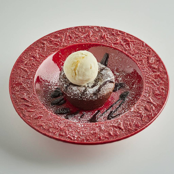 Un dessert una torta di lava al cioccolato condita con una pallina di gelato alla vaniglia, guarnita con una foglia di menta, un fiore commestibile e un frutto della physalis, il tutto servito su un piatto di ceramica rossa brillante. - Foto, immagini