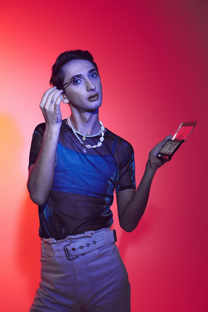 соблазнительный гей в повседневной одежде, используя палитру теней для век и глядя на камеру на ярком фоне - Фото, изображение