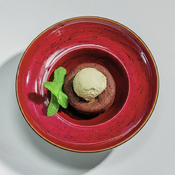 Ein Dessert, ein Schokoladen-Lavakuchen mit einer Kugel Vanilleeis, garniert mit einem Minzblatt, einer essbaren Blume und einer Physalis-Frucht, serviert auf einem leuchtend roten Keramikteller. - Foto, Bild