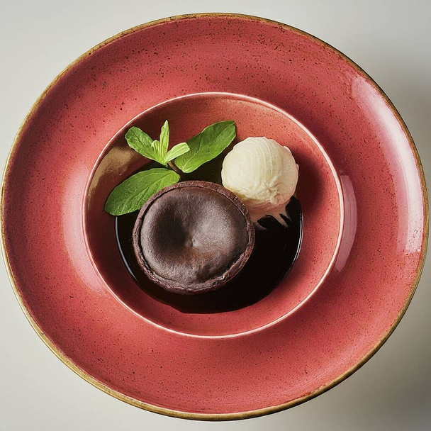 Een dessert, een chocoladetaart met een bolletje vanille-ijs, versierd met een muntblad, een eetbare bloem en een fysalis-vrucht, geserveerd op een rode keramische plaat.. - Foto, afbeelding