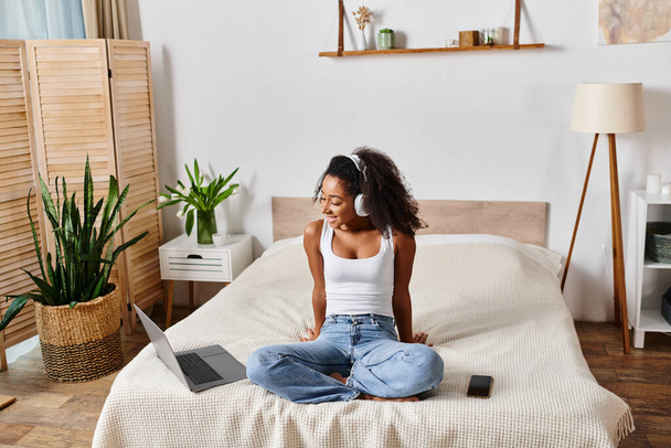 Μια σγουρή Αφρο-Αμερικανίδα με ένα μπλουζάκι κάθεται σε ένα κρεβάτι, βυθισμένη στο λάπτοπ της.. - Φωτογραφία, εικόνα