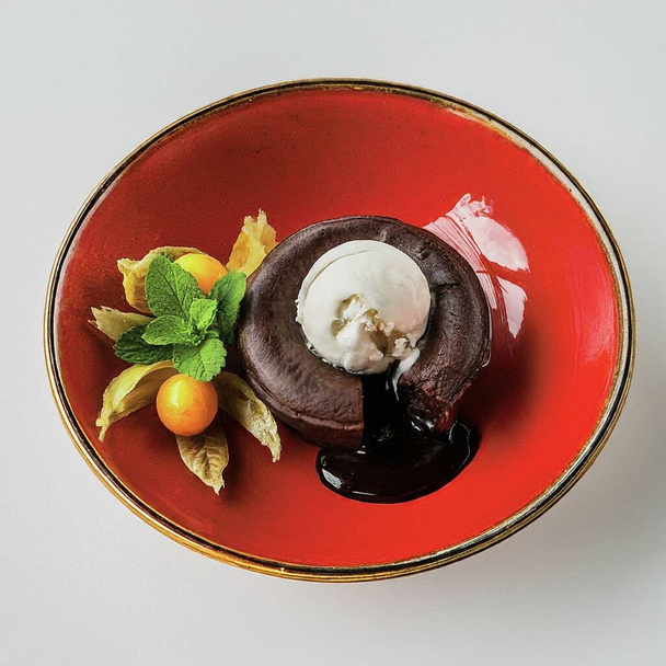 Un dessert una torta di lava al cioccolato condita con una pallina di gelato alla vaniglia, guarnita con una foglia di menta, un fiore commestibile e un frutto della physalis, il tutto servito su un piatto di ceramica rossa brillante. - Foto, immagini
