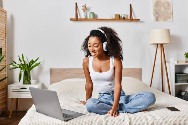 Askılı tişörtlü kıvırcık Afrikalı Amerikalı bir kadın modern bir yatak odasında dizüstü bilgisayarına odaklanmış bir şekilde bir yatakta oturuyor.. - Fotoğraf, Görsel