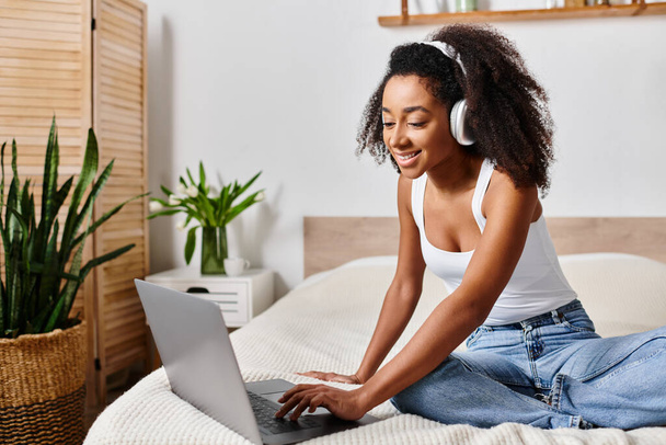 Femme afro-américaine bouclée dans un débardeur, assise sur un lit, intensément concentrée sur l'utilisation d'un ordinateur portable dans une chambre moderne. - Photo, image