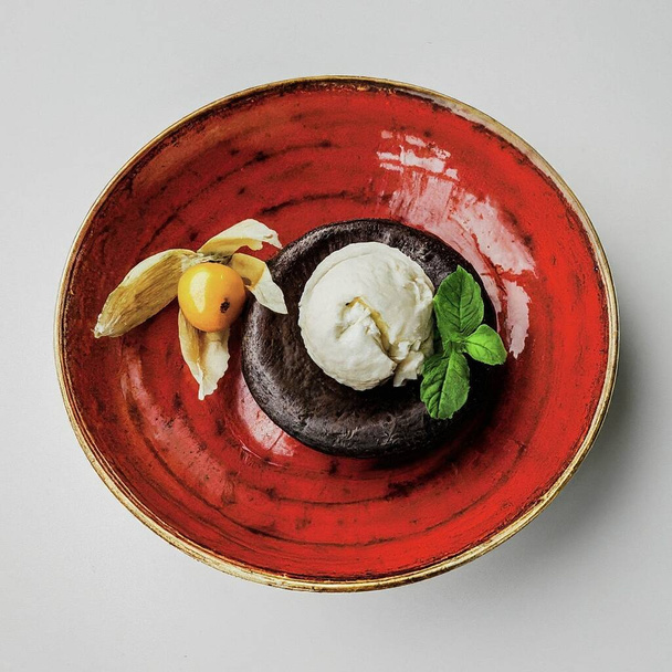 Dezert čokoládový lávový dort zakončený kopečkem vanilkové zmrzliny, zdobený mátovým listem, jedlou květinou a fyzalisovým ovocem, vše podávané na lesklém červeném keramickém talíři. - Fotografie, Obrázek