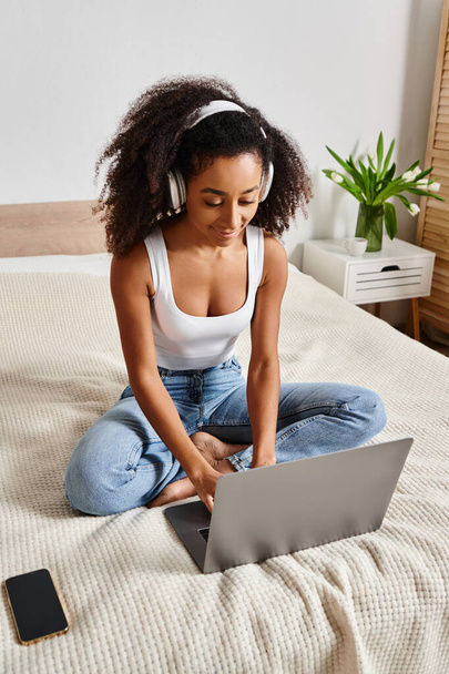 Mujer afroamericana rizada sentada en la cama, escribiendo en el ordenador portátil en un entorno de dormitorio moderno. - Foto, imagen